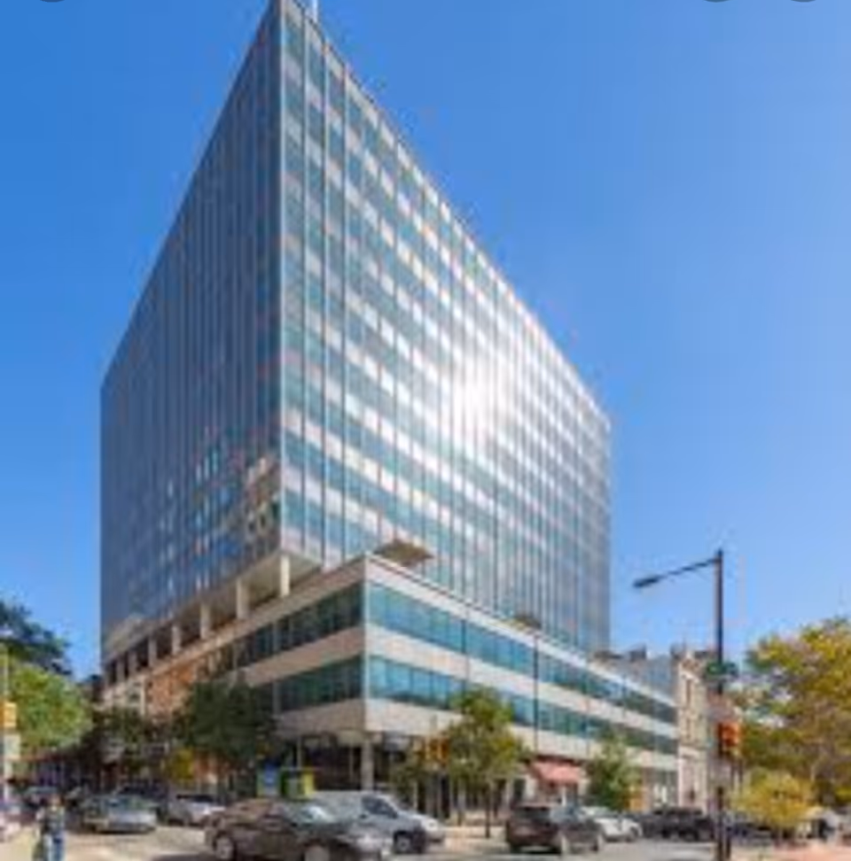Tall glass building on a city street with parked cars and trees.