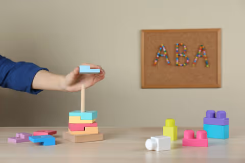 Hand stacks colorful blocks on a table in an ABA therapy room.