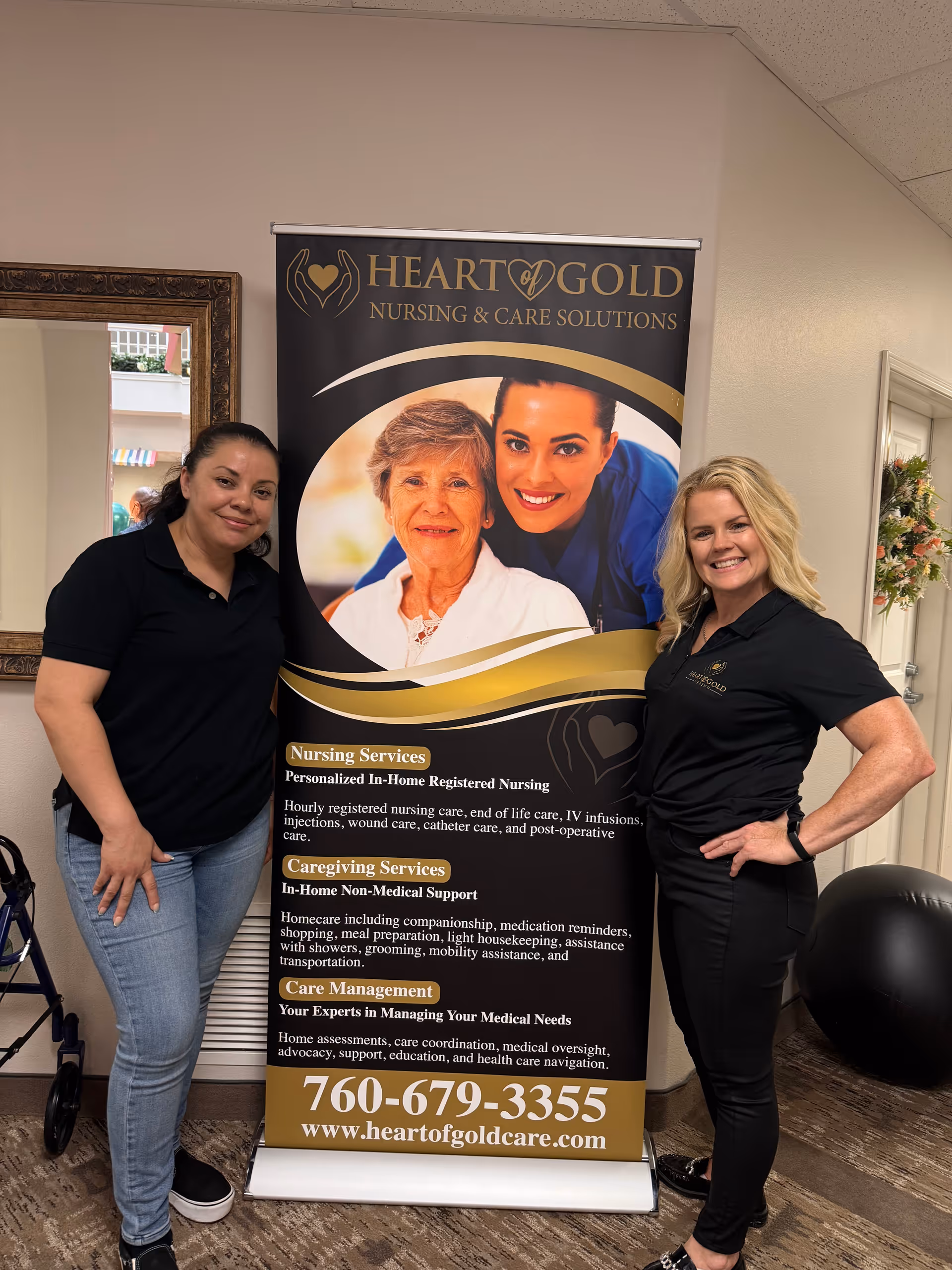 Two women pose beside a Heart of Gold Nursing banner in an interior hallway.