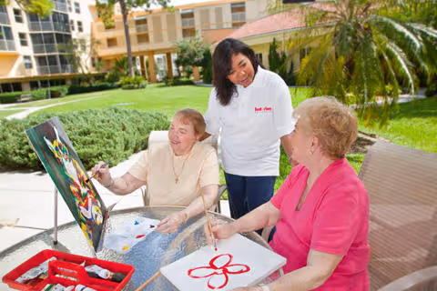 Three seniors painting outdoors on a sunny patio, assisted by a caregiver.