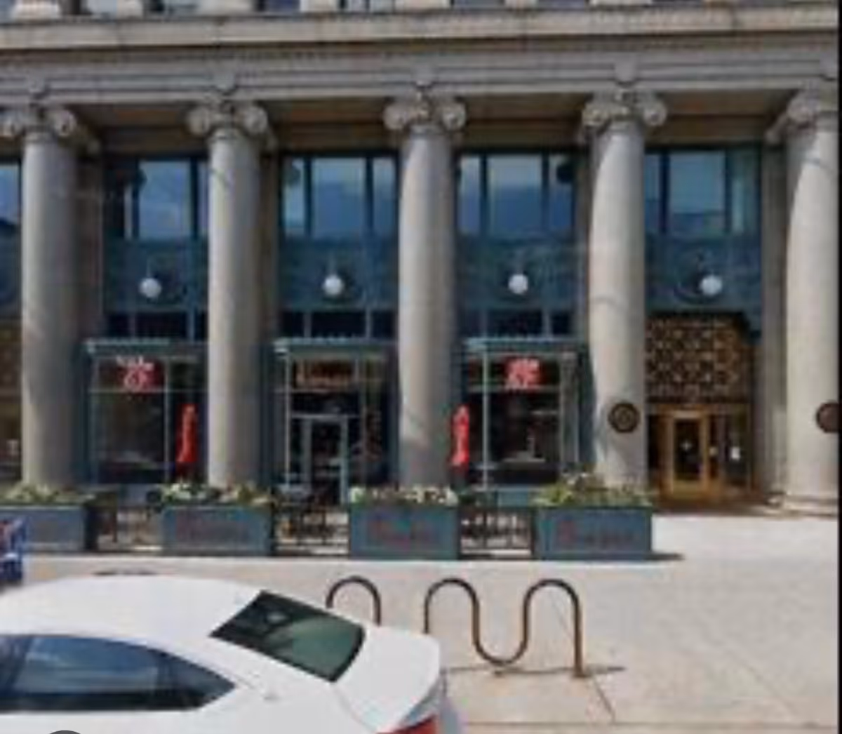 Front of a large building with tall columns and glass doors, cars parked outside.