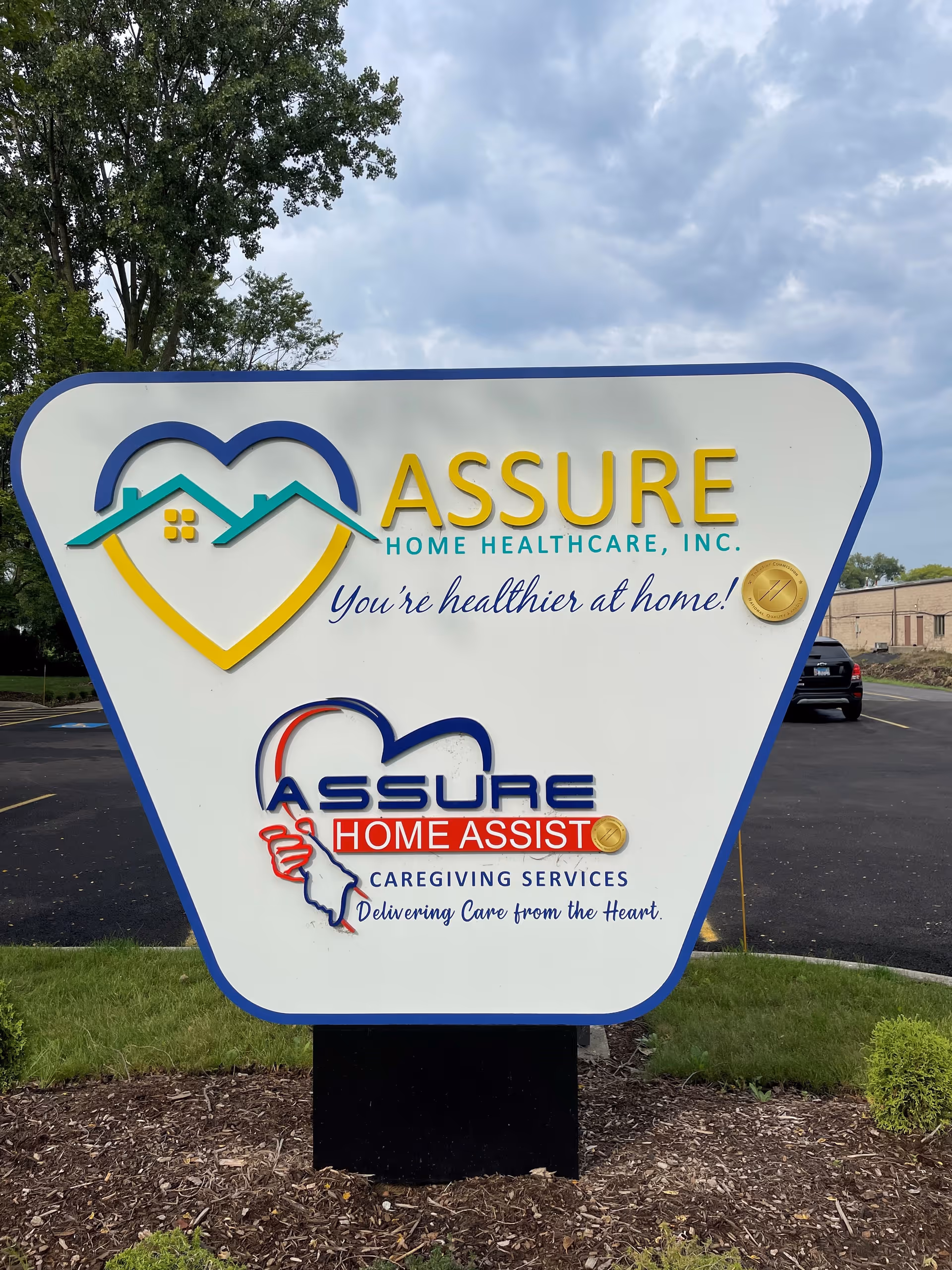 Large outdoor building-front sign advertising Assure Home Healthcare with heart logos and motto.