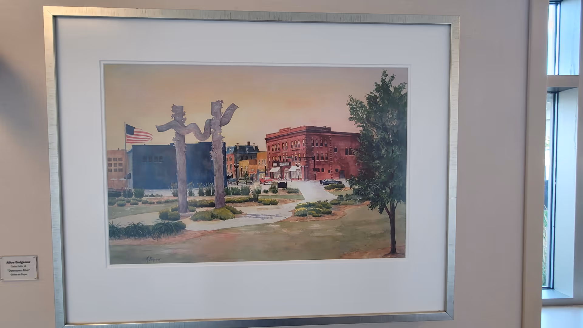 Framed painting of a town square hangs on a pale interior wall.