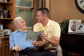 An elderly man and a caregiver chat while looking at a pamphlet in a living room.