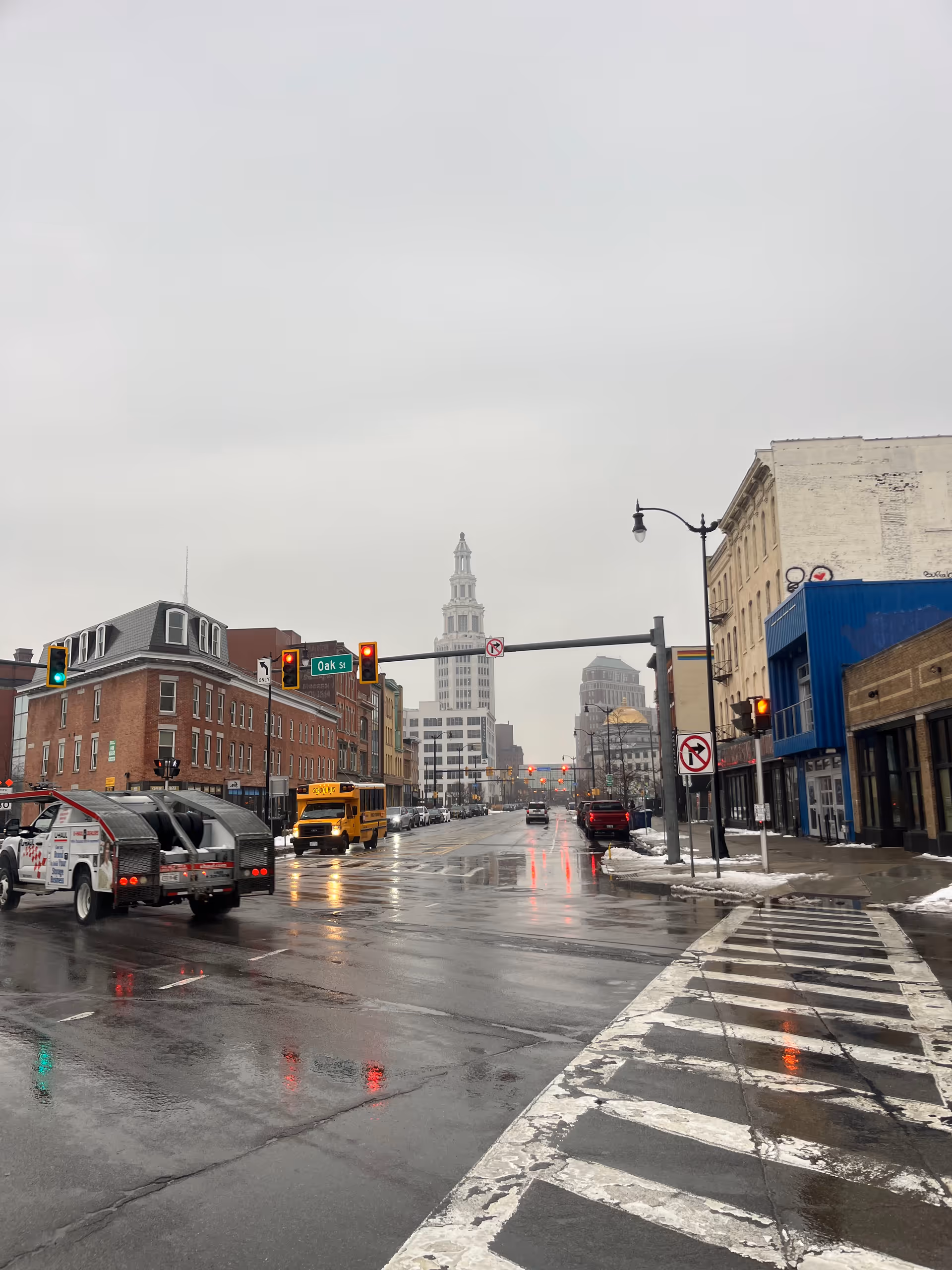 Urban rainy street with wet pavement, traffic signals, and storefronts along a downtown avenue.