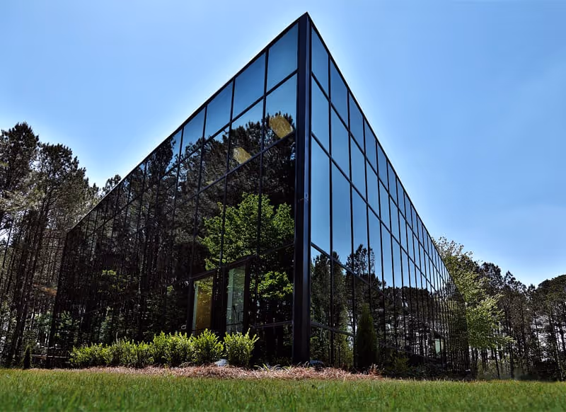 Modern glass-front building reflects surrounding trees, set in a grassy landscape under a clear blue sky.