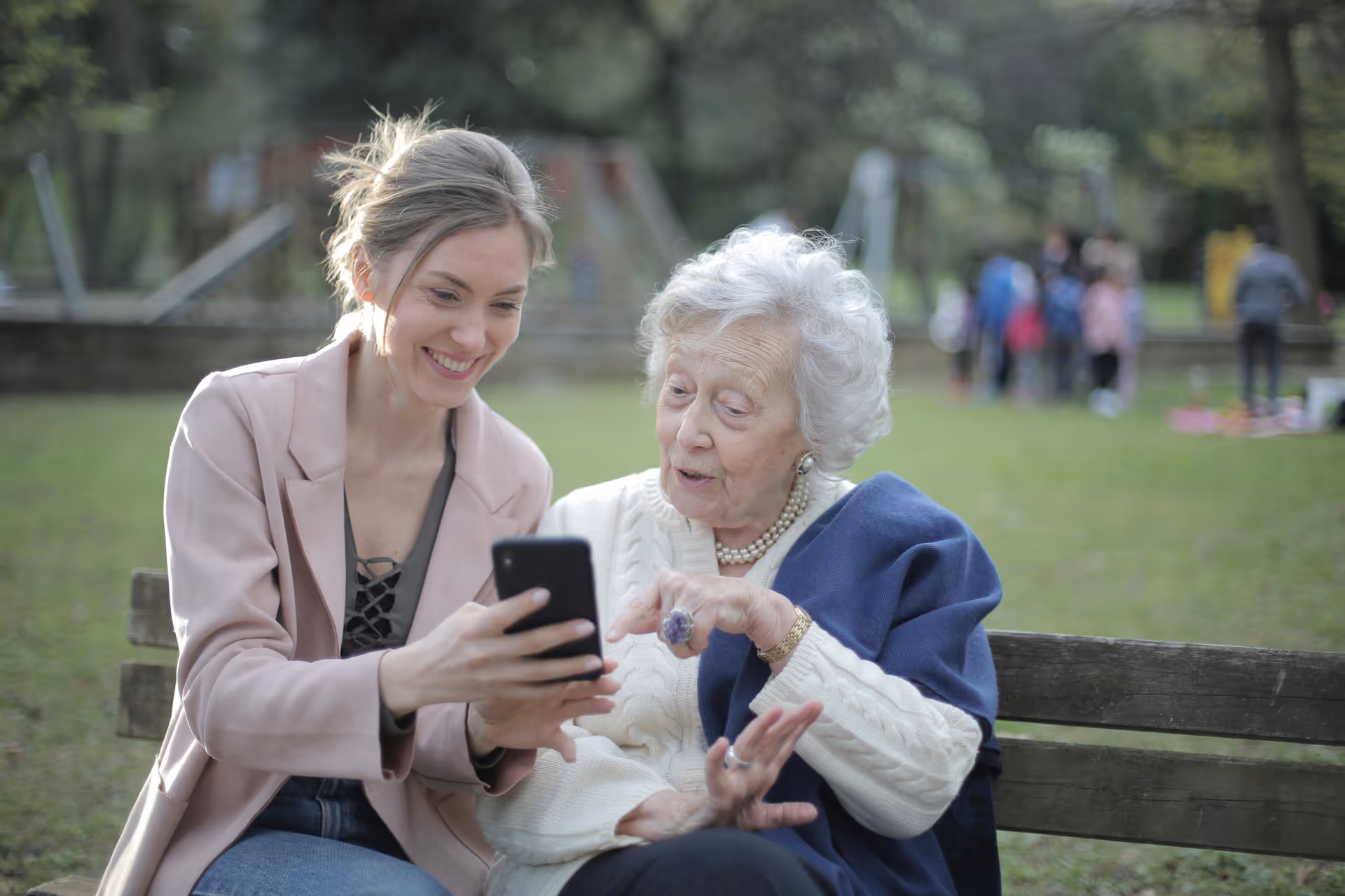 Two women, a caregiver and an elderly resident, sit on a park bench using a smartphone.