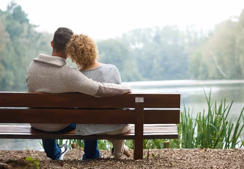 Two people sit on a wooden bench beside a lake in a park setting.