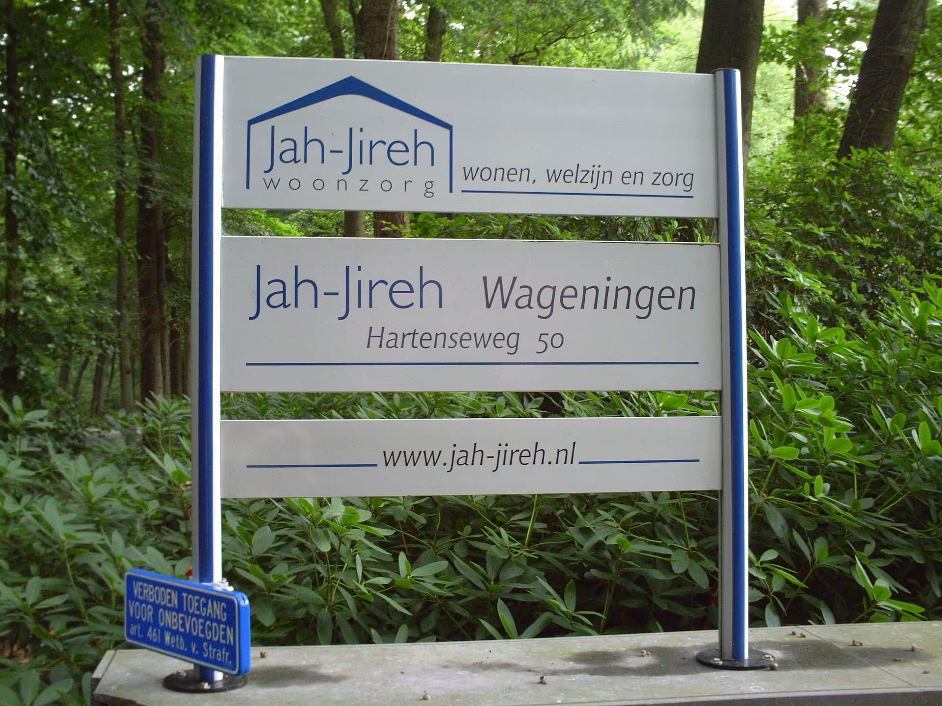 Outdoor sign for Jah-Jireh Woonzorg facility amid green trees and shrubs.