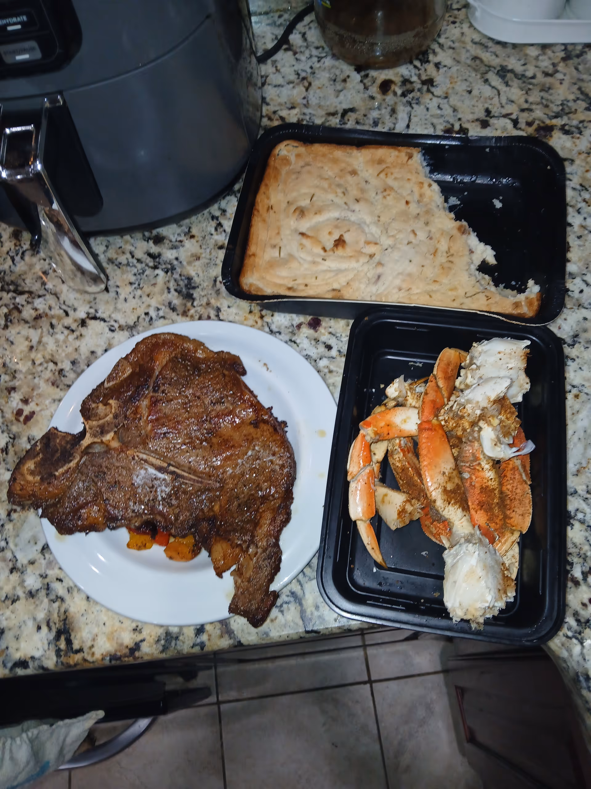 A granite kitchen counter with cooked steak, crab legs, and sliced bread in trays.